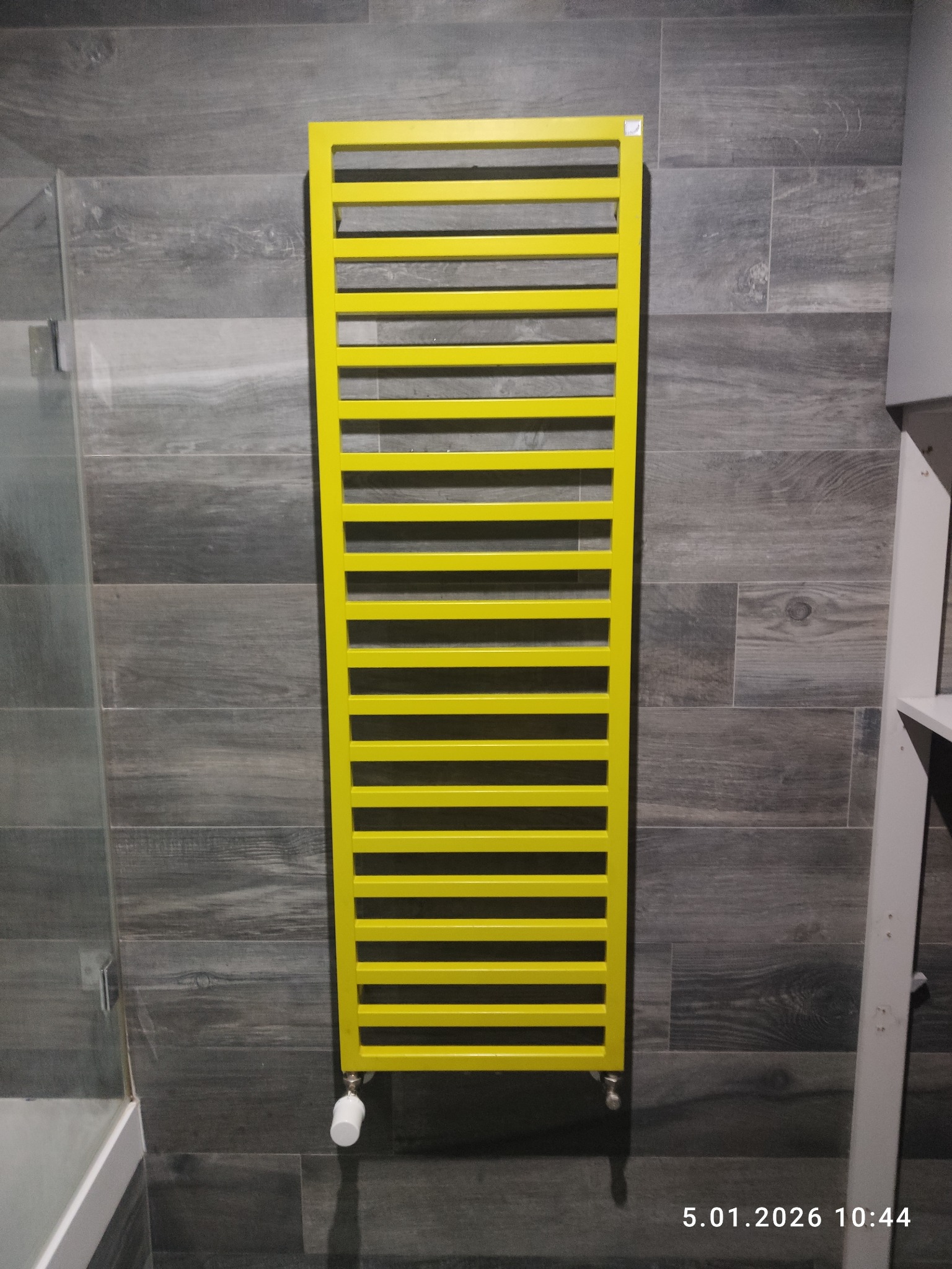 Żółty, drabinkowy grzejnik łazienkowy zamontowany na szarej ścianie z płytek imitujących drewno. Widoczny fragment kabiny prysznicowej i biały element wyposażenia.