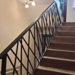 Wewnętrzne schody z drewnianymi stopniami i czarną metalową poręczą o wzorze romboidalnym, oświetlone lampą sufitową.