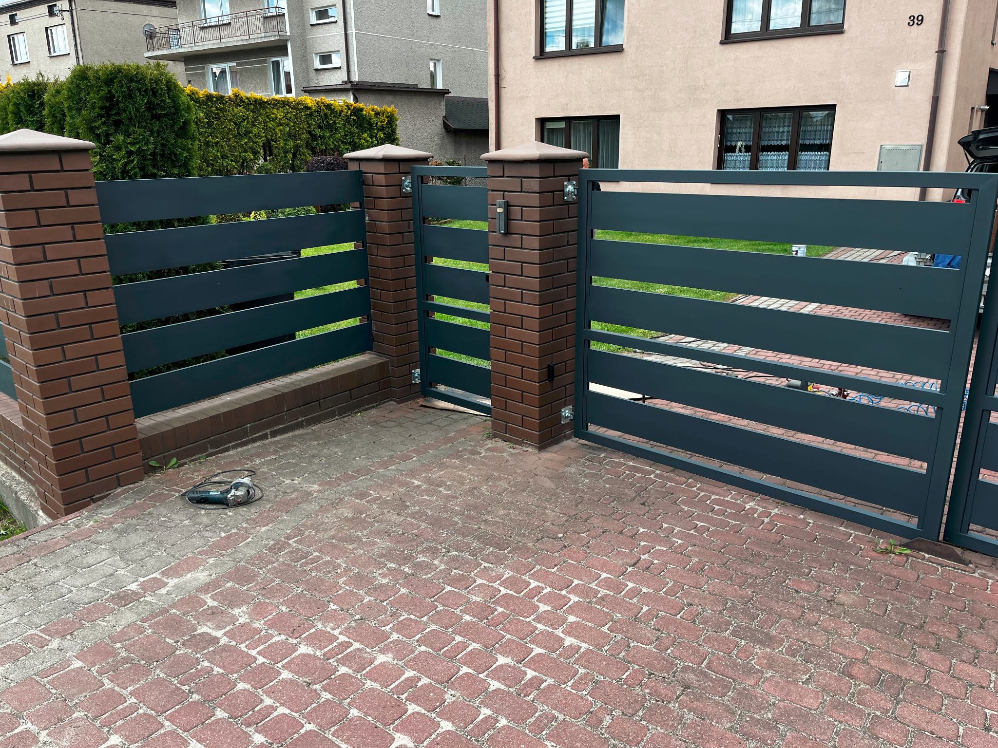Nowoczesna brama wjazdowa z poziomymi panelami w kolorze grafitowym, osadzona na ceglanych słupkach, z widoczną kostką brukową i szlifierką kątową obok słupka.