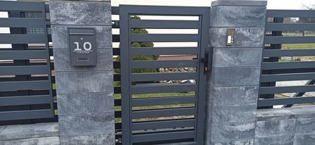 Nowoczesna brama wjazdowa z poziomymi przęsłami, osadzona między murowanymi słupkami z szarego betonu, jeden z nich z zamontowaną skrzynką na listy z numerem 10.