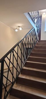 Wewnętrzne schody z drewnianymi stopniami i czarną metalową poręczą o wzorze romboidalnym, oświetlone lampą sufitową.