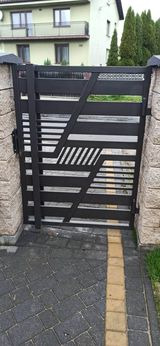 Nowoczesna brama wjazdowa z poziomymi, czarnymi panelami i geometrycznym wzorem, osadzona między kamiennymi słupkami na tle domu.