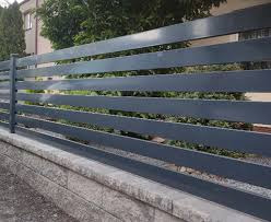 Nowoczesna, pozioma poręcz/ogrodzenie w kolorze antracytowym, zamontowana na niskim murku z szarej kostki, z zielenią w tle.