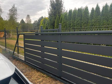 Nowoczesna, przesuwna brama wjazdowa w kolorze grafitowym, z poziomymi szczelinami, zamontowana na posesji z widocznym ogrodzeniem z siatki i tujami w tle.