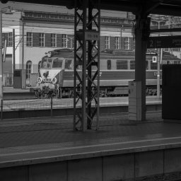 Adrian Trzaska - Czarno-białe zdjęcie lokomotywy EP07-361 na peronie kolejowym, widok zza metalowej konstrukcji wsporczej z numerem 159-19 N, budynek w tle.