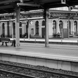 Adrian Trzaska - Czarno-białe zdjęcie peronu kolejowego z osobami siedzącymi na ławkach w tle, widoczne tory i elementy konstrukcyjne zadaszenia.