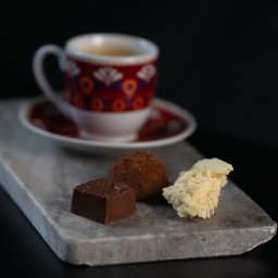 Adrian Trzaska - Kompozycja z filiżanki kawy z wzorzystym motywem, czekoladowego kwadratu z wytłoczonym ziarnem kawy, trufli i białej czekolady na marmurowej podstawce, na czarnym tle.