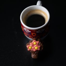 Adrian Trzaska - Filiżanka kawy z wzorzystym motywem i czekoladowy bukiecik na czarnym tle, ujęcie z góry.