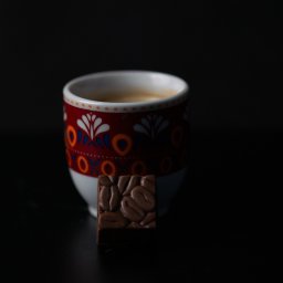 Adrian Trzaska - Espresso w filiżance z folkowym wzorem i czekoladka z wytłoczonymi ziarnami kawy na czarnym tle.