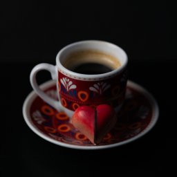Adrian Trzaska - Filiżanka espresso z wzorzystym zdobieniem i czekoladka w kształcie serca na spodku, na czarnym tle.