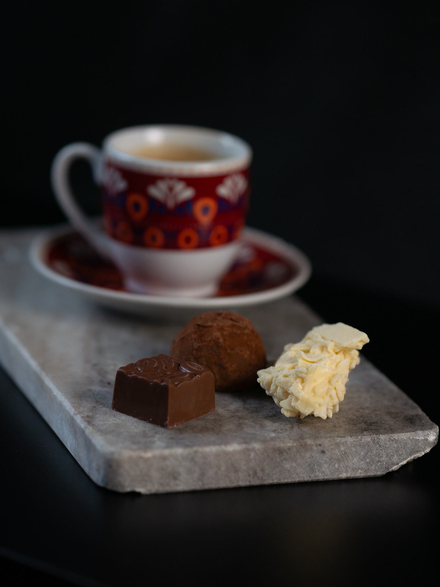 Kompozycja z filiżanki kawy z wzorzystym motywem, czekoladowego kwadratu z wytłoczonym ziarnem kawy, trufli i białej czekolady na marmurowej podstawce, na czarnym tle.