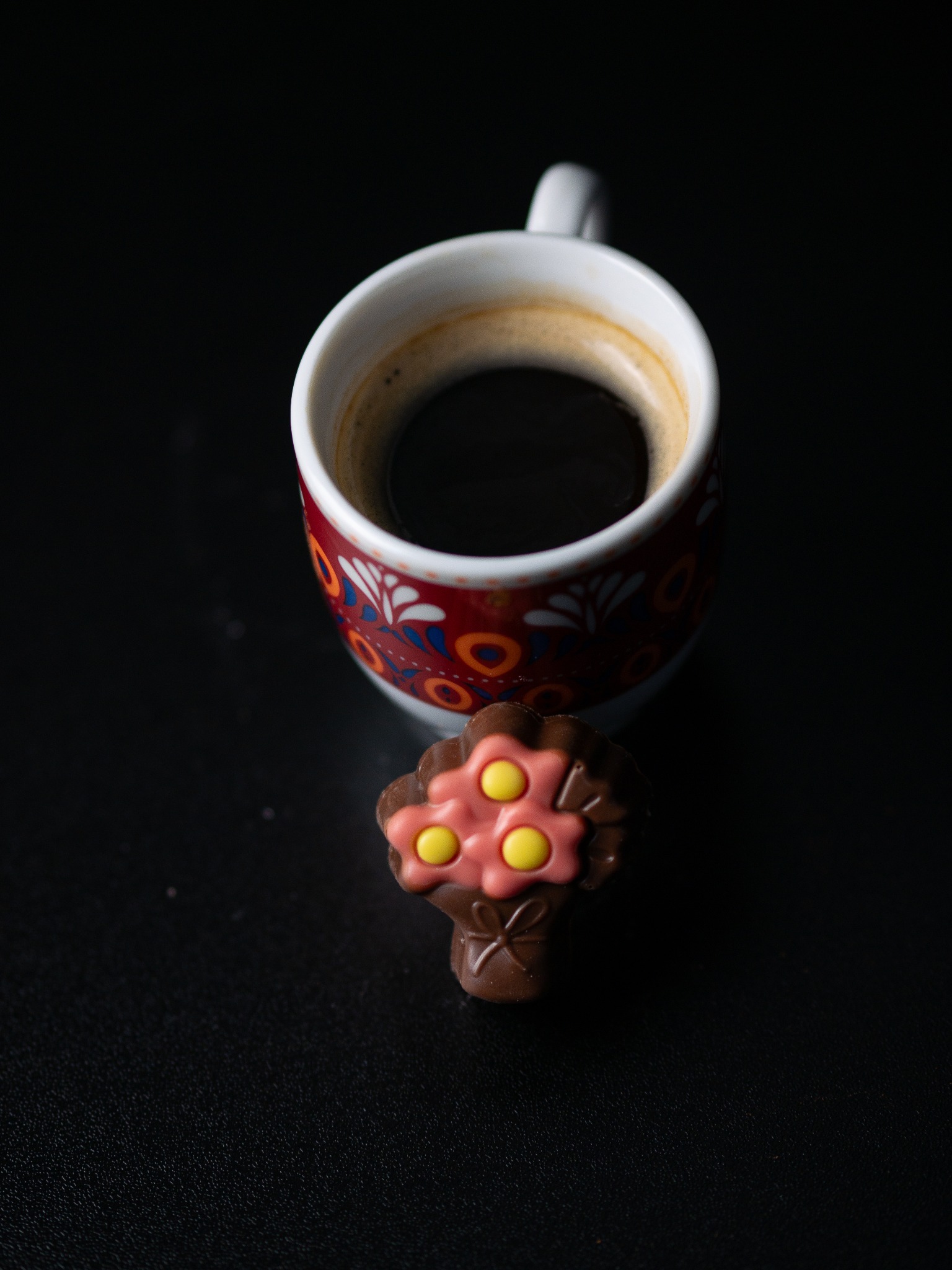 Filiżanka kawy z wzorzystym motywem i czekoladowy bukiecik na czarnym tle, ujęcie z góry.