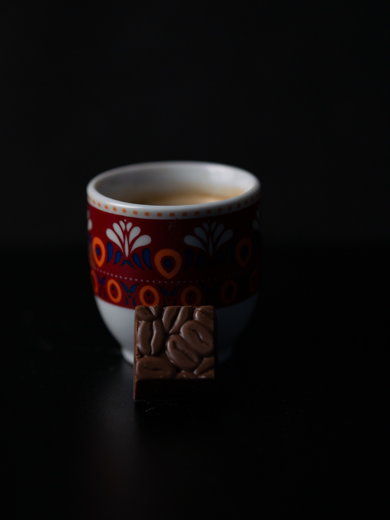 Espresso w filiżance z folkowym wzorem i czekoladka z wytłoczonymi ziarnami kawy na czarnym tle.