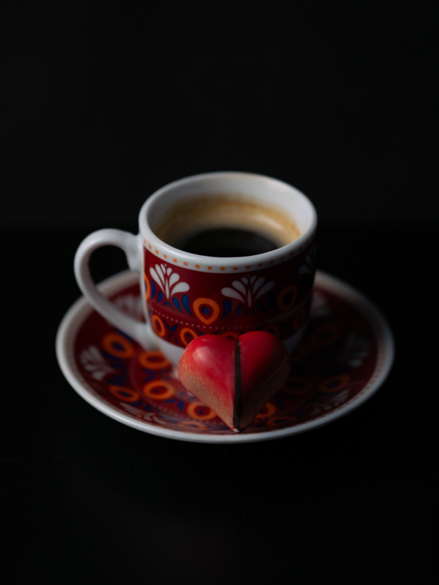 Filiżanka espresso z wzorzystym zdobieniem i czekoladka w kształcie serca na spodku, na czarnym tle.