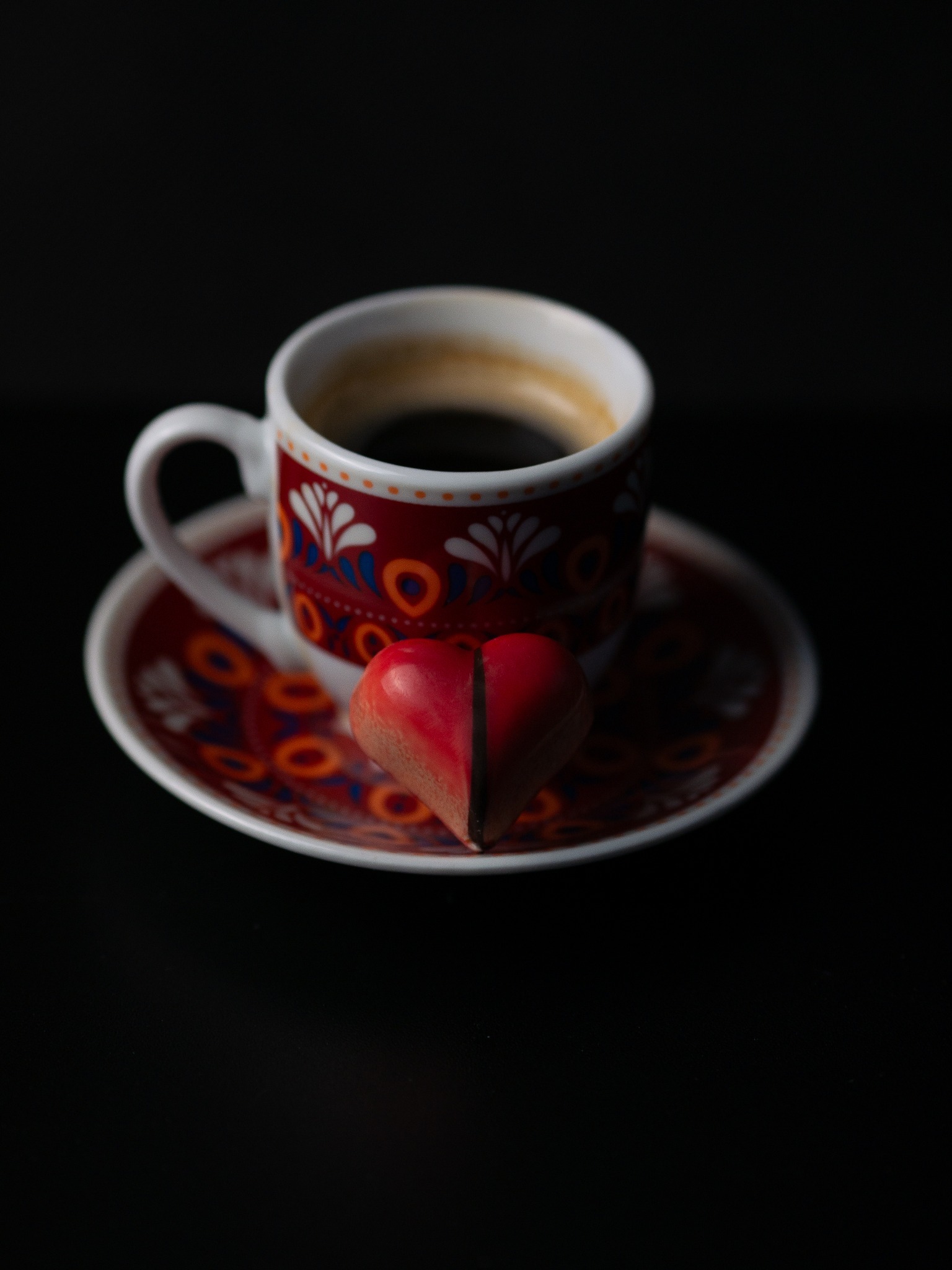 Czerwony cukierek w kształcie serca oparty o filiżankę espresso na spodku z folkowym wzorem na czarnym tle.
