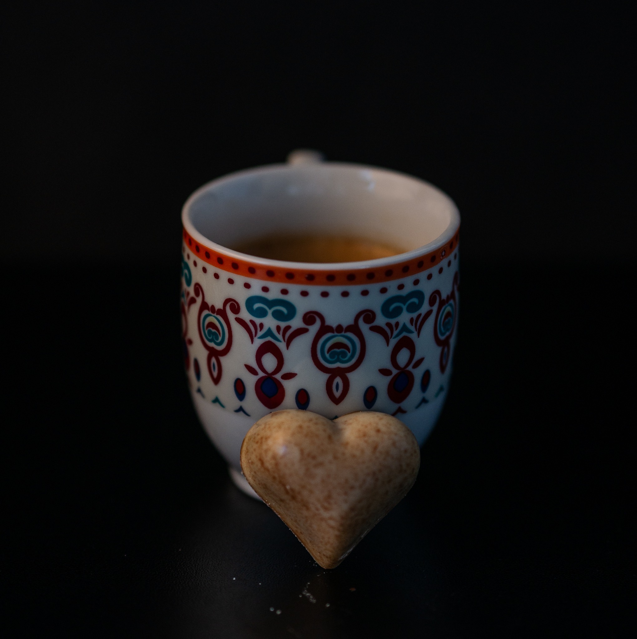 Filiżanka z wzorzystym motywem etnicznym, wypełniona kawą, obok której leży czekoladka w kształcie serca na czarnym tle.