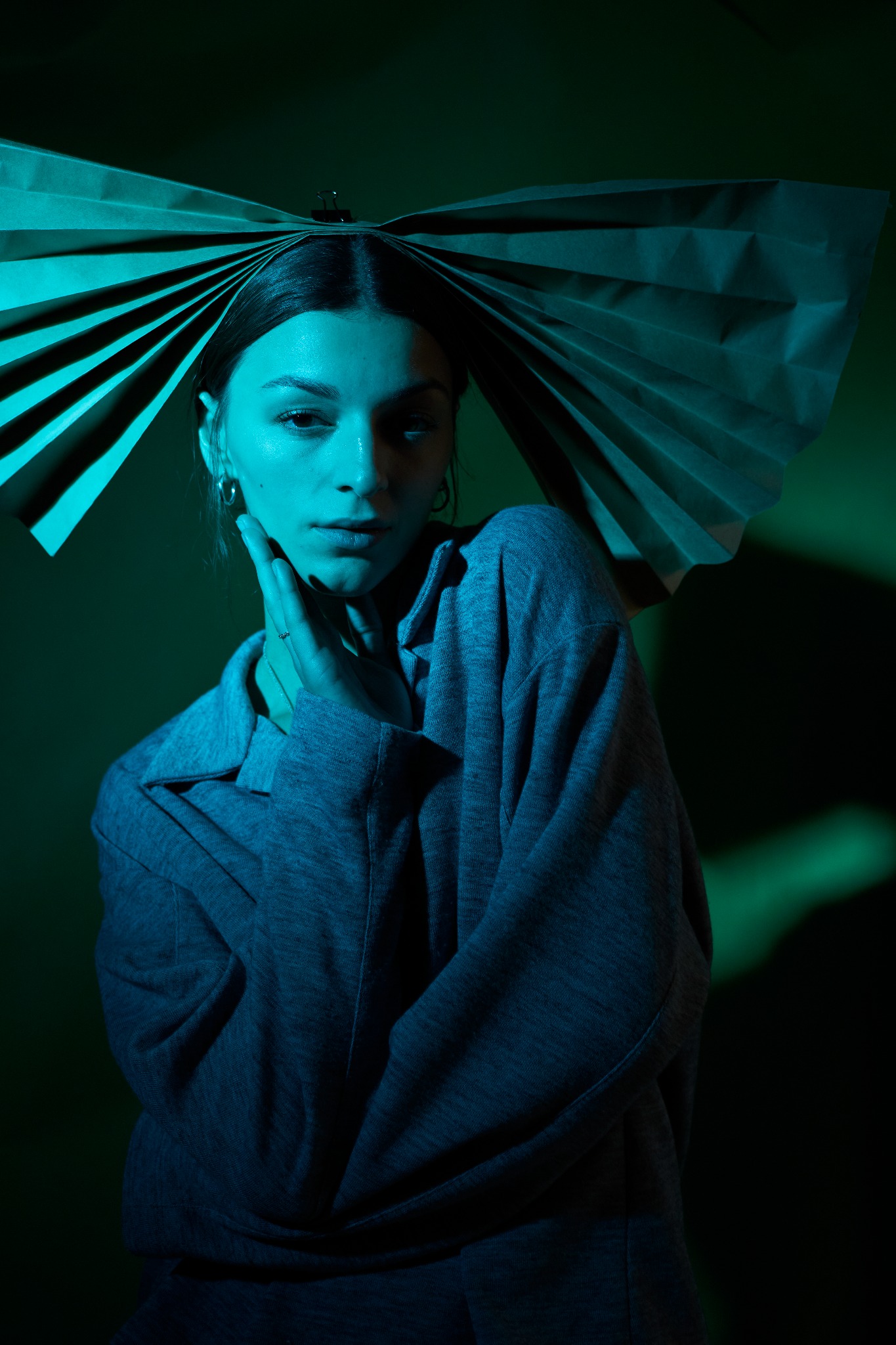 Portret kobiety w niebiesko-zielonym oświetleniu, z papierową ozdobą na głowie, dotykającej szyi.