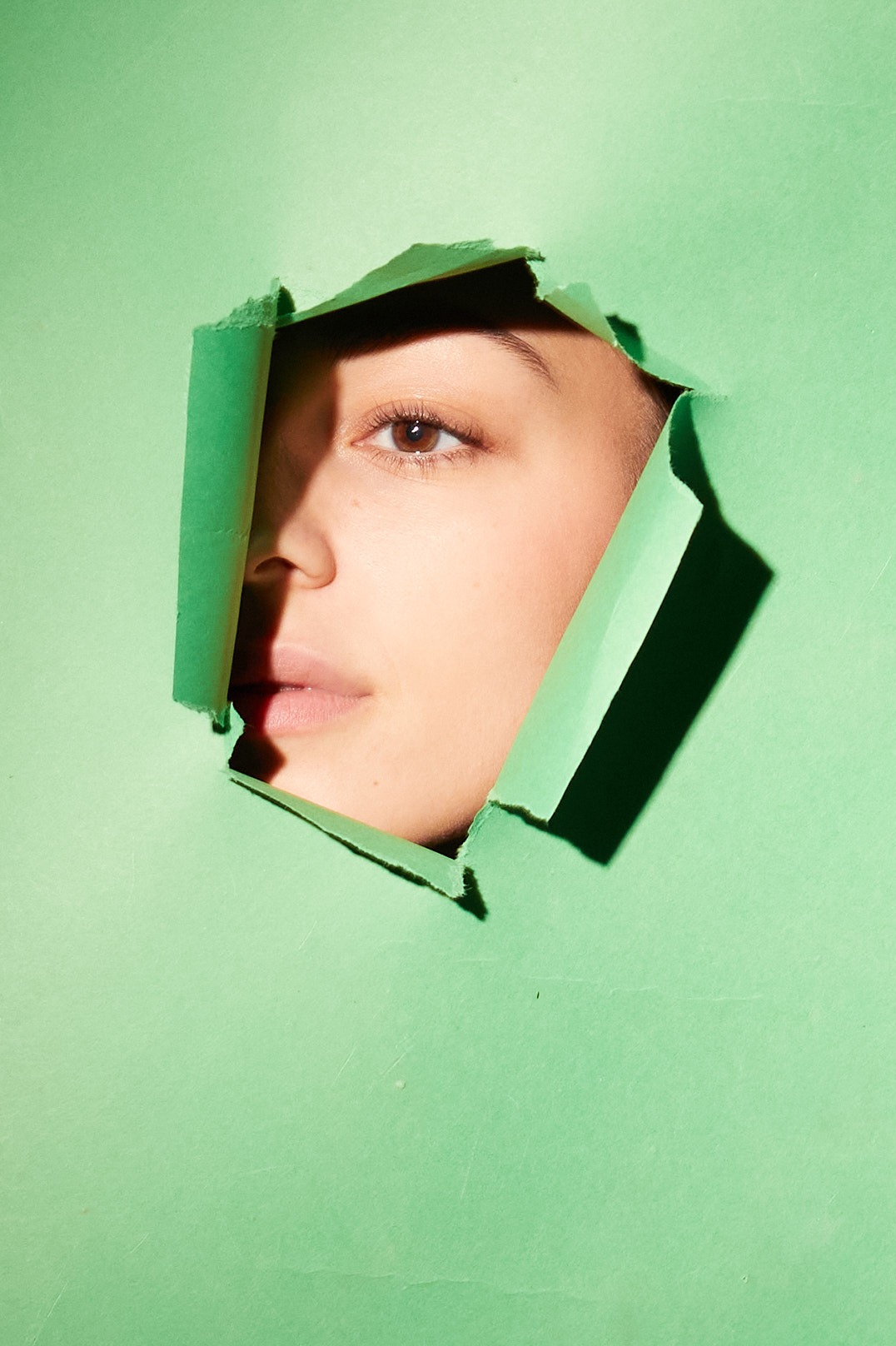 Portret kobiety, której część twarzy widoczna jest przez dziurę w zielonym papierze, z wyraźnym cieniem na policzku i intensywnym spojrzeniem.