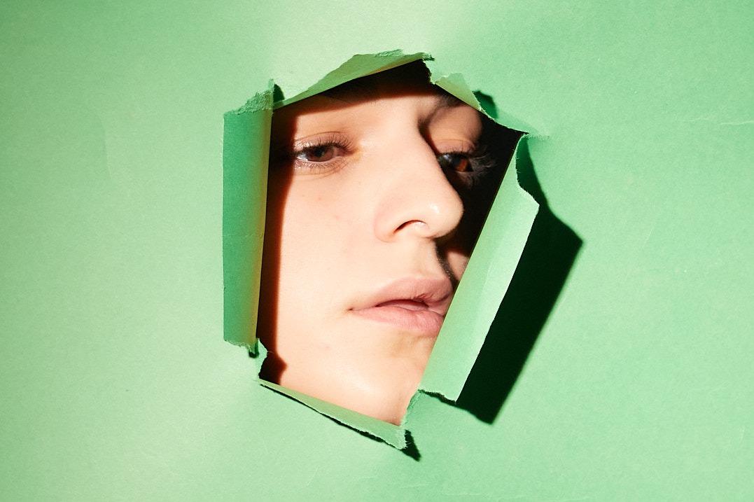 Portret kobiety spoglądającej przez dziurę w zielonym papierze, z cieniem padającym na prawą stronę twarzy i papieru.