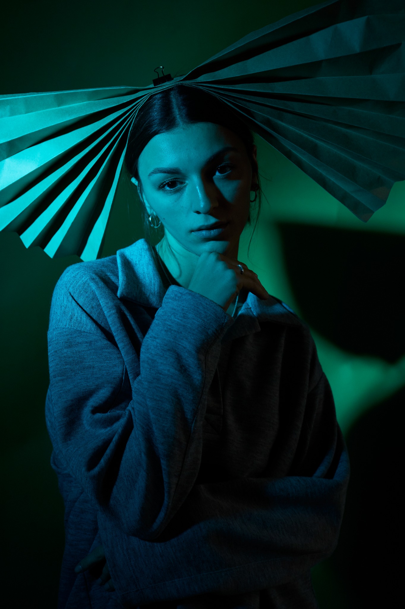Portret kobiety z papierową ozdobą na głowie, oświetlony zielono-niebieskim światłem, z ręką pod brodą i pierścionkiem na palcu.