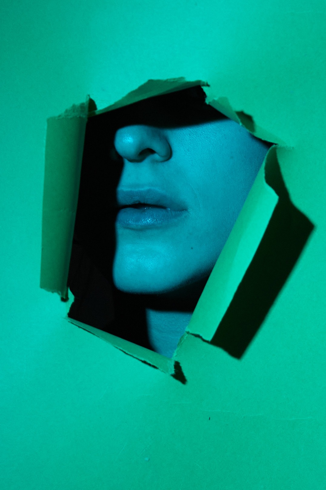 Fragment twarzy modelki, usta i nos, wyłaniający się z otworu w zielonym papierze, oświetlony niebieskim światłem w Bydgoszczy.