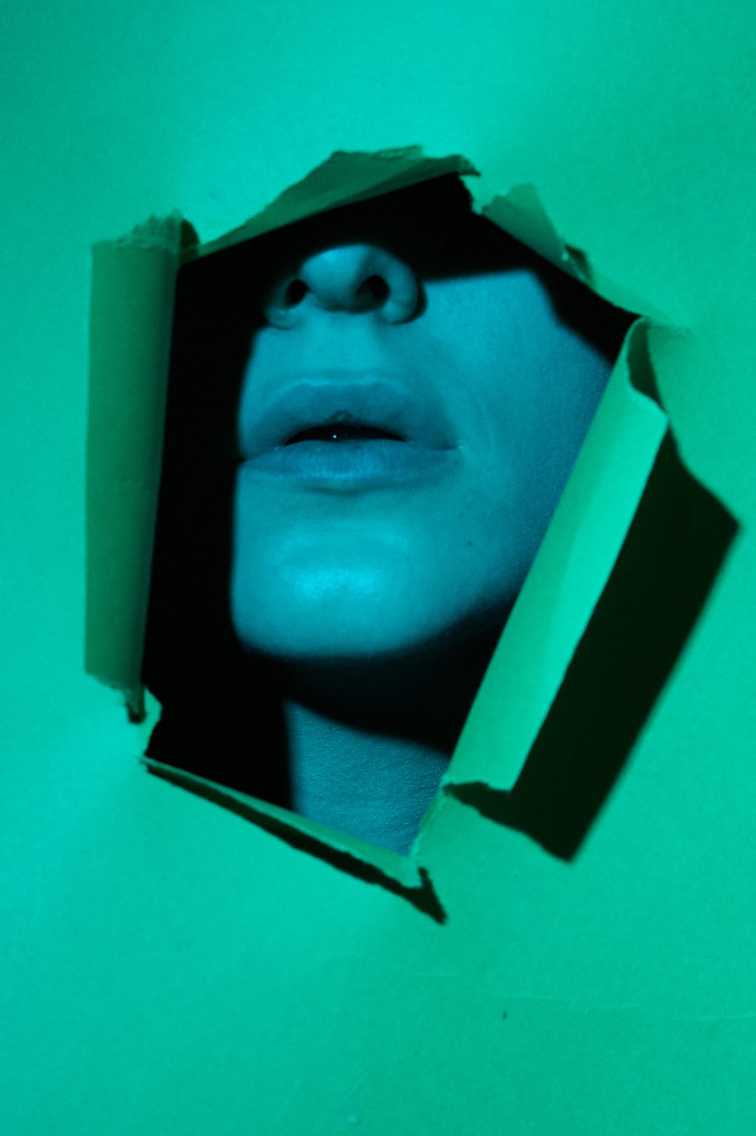Fragment twarzy modelki widoczny przez podarty otwór w zielonej kartce, oświetlony chłodnym niebieskim światłem.
