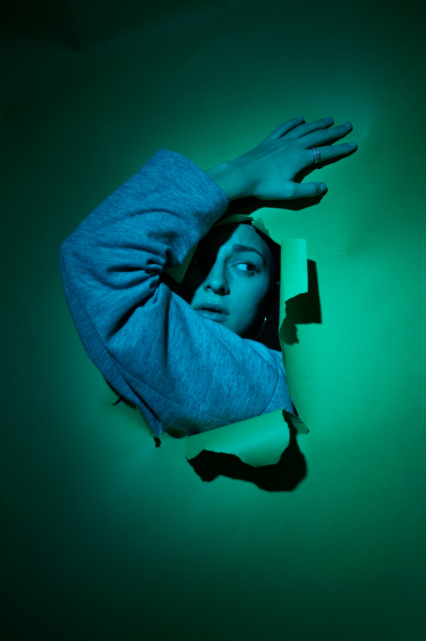 Portret kobiety w szarej bluzie, spoglądającej w prawo przez podarte zielone tło, oświetlony zielonym światłem.