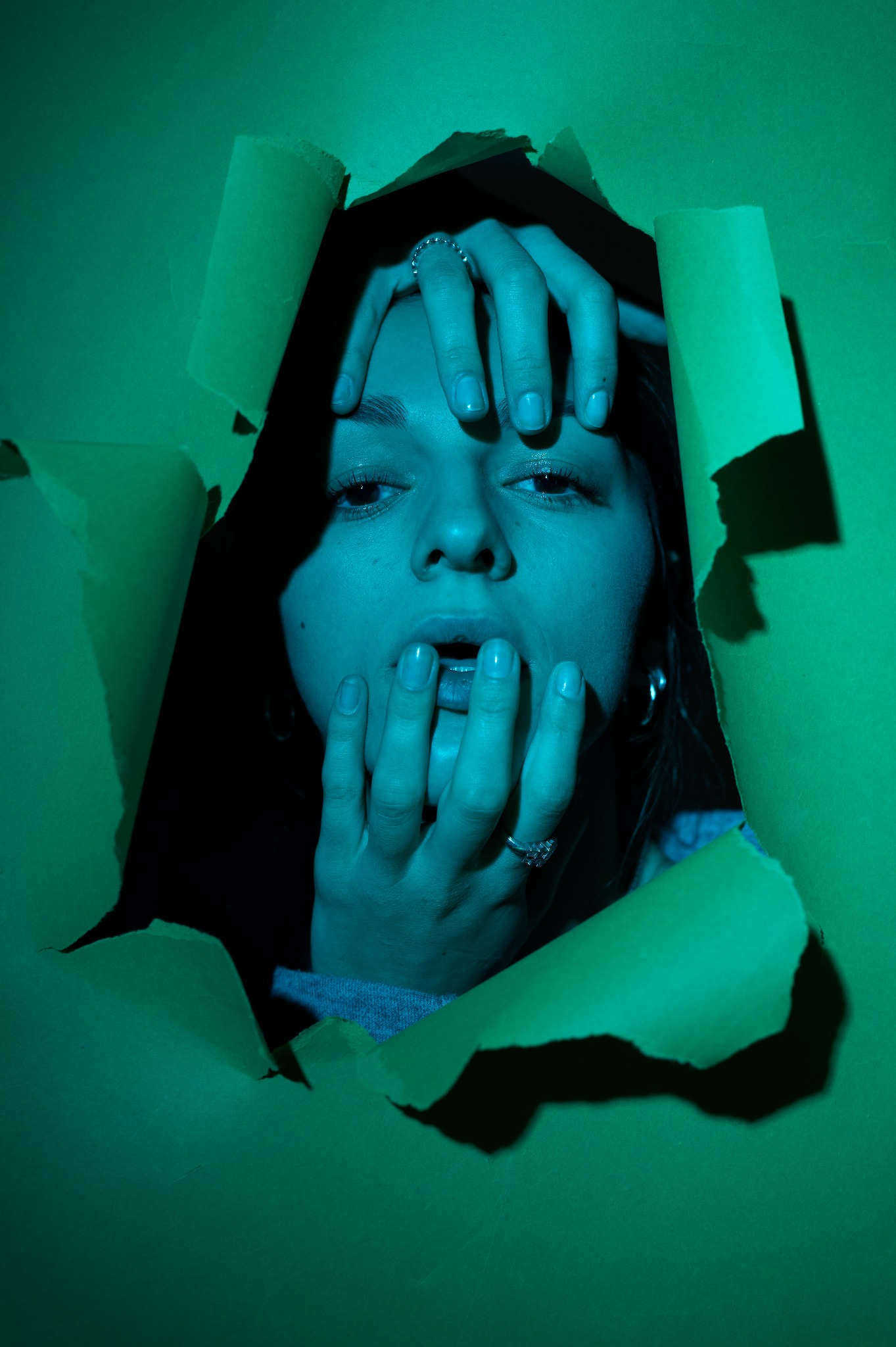 Portret kobiety z pierścionkami, spoglądającej przez rozdarte, zielone tło, oświetlony turkusowym światłem.