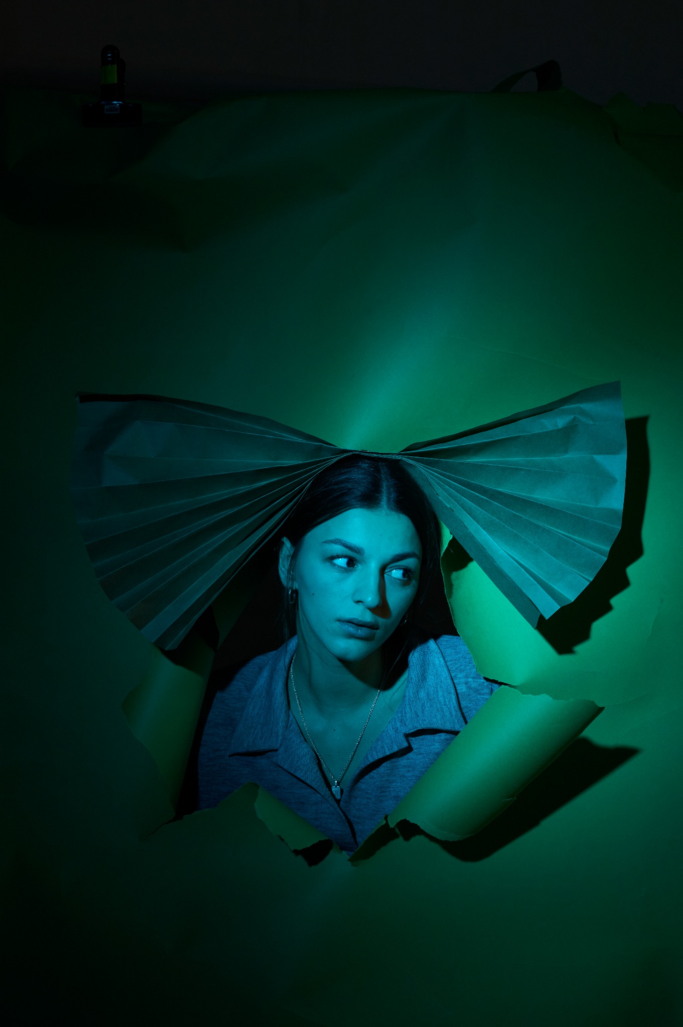 Portret kobiety spoglądającej w bok, wyłaniającej się z rozdartego zielonego tła, z papierową kokardą nad głową, oświetlony niebiesko-zielonym światłem.