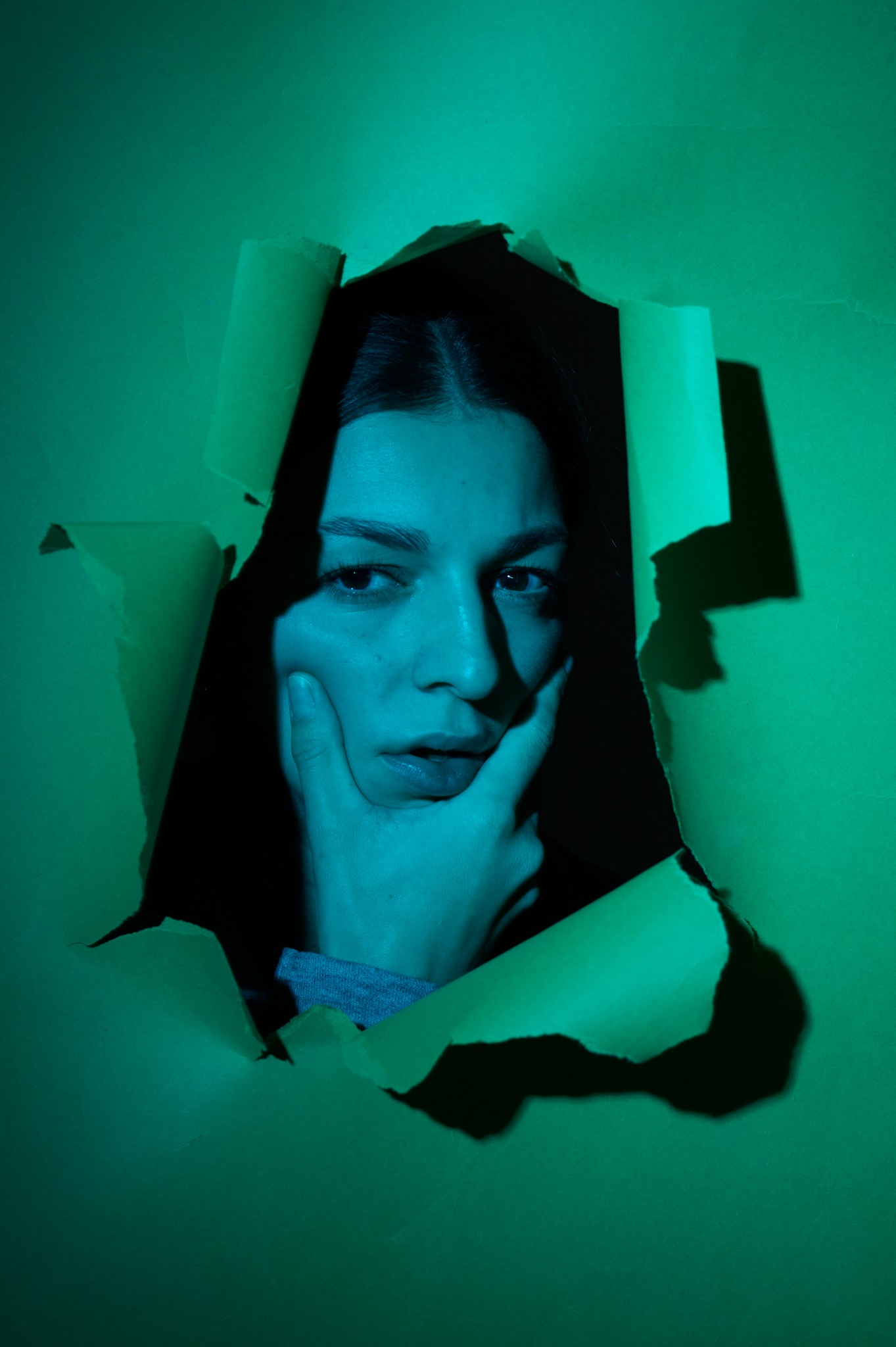 Portret kobiety spoglądającej w bok, wyłaniającej się z podartego, zielonego papieru, oświetlonej turkusowym światłem.