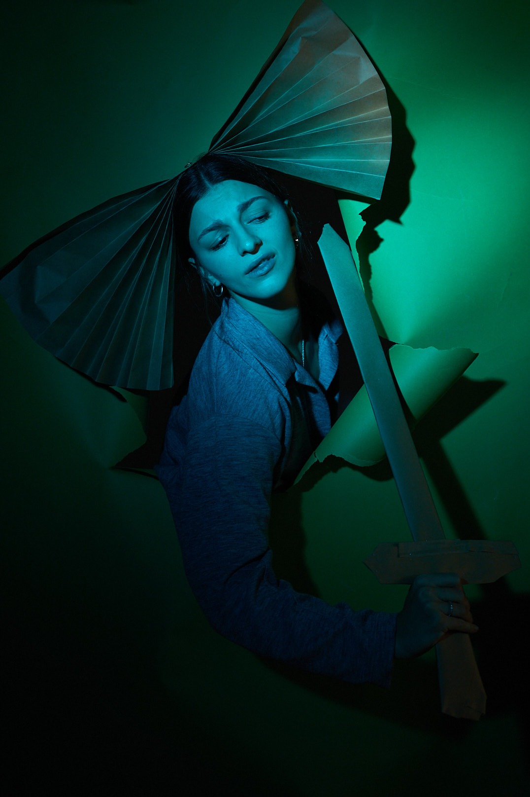 Portret kobiety wyłaniającej się z dziury w zielonym papierze, trzymającej tekturowy miecz, z papierowym wachlarzem nad głową, oświetlony chłodnym, zielono-niebieskim światłem.