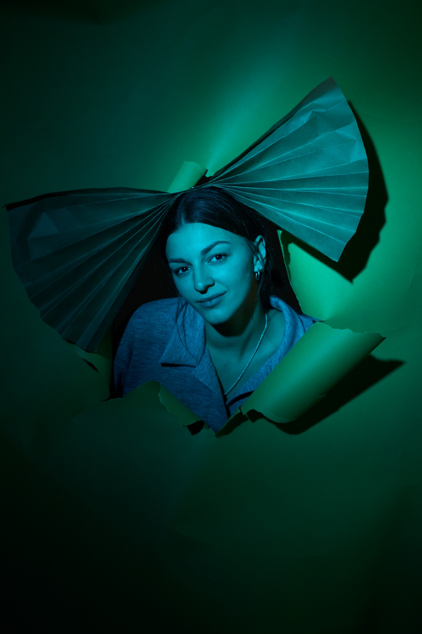 Portret kobiety spoglądającej przez dziurę w zielonym papierze, z papierowym wachlarzem nad głową, oświetlony turkusowym światłem.