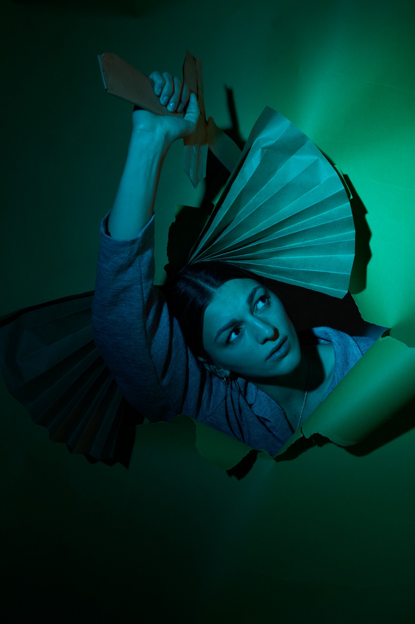 Kobieta spogląda w górę, wystając z rozdartego zielonego papieru, trzymając w dłoni tekturowy rekwizyt, oświetlona zielonym światłem.