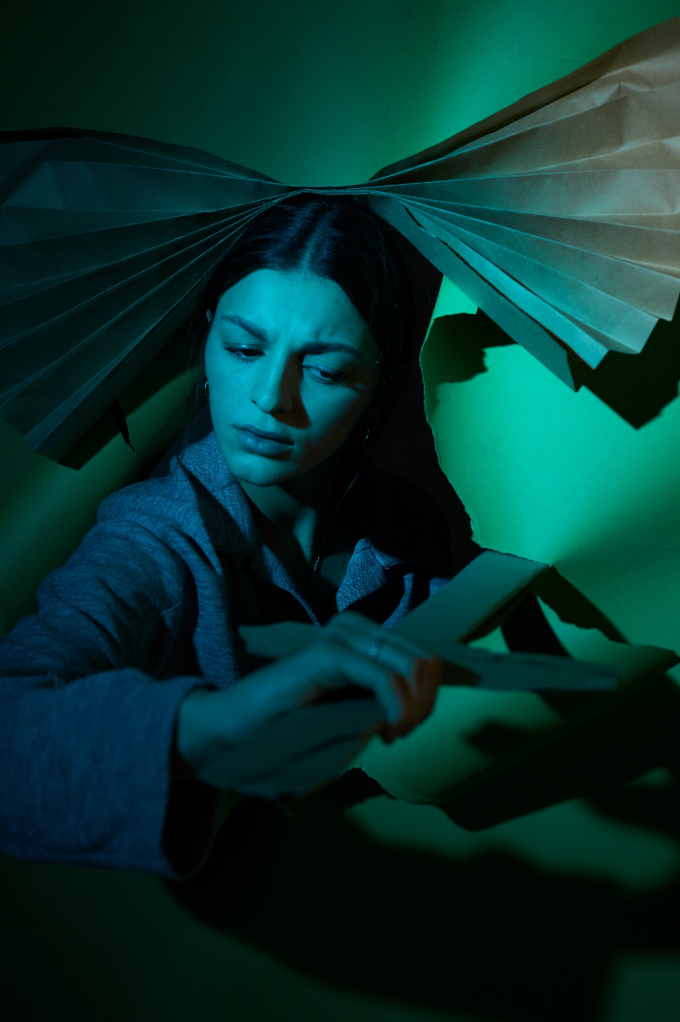 Portret kobiety w niebiesko-zielonym oświetleniu, otoczonej papierowymi elementami, tworzącymi abstrakcyjną kompozycję nad głową i w dłoniach.