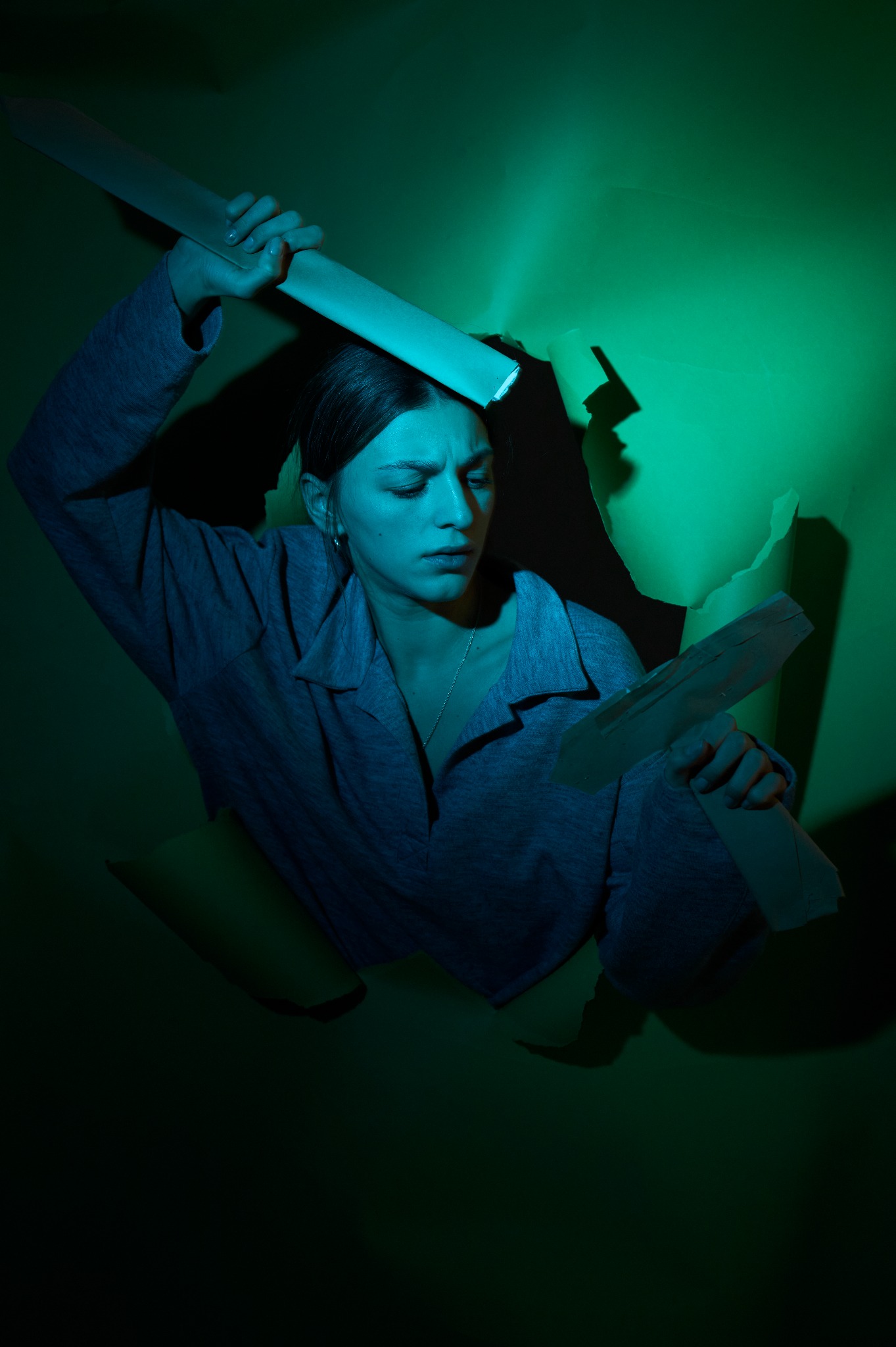 Kobieta w szarej koszuli patrzy w dół, wystając z dziury w zielonym papierze, trzymając kawałki podartych tektur w dłoniach, oświetlona zielonym światłem.