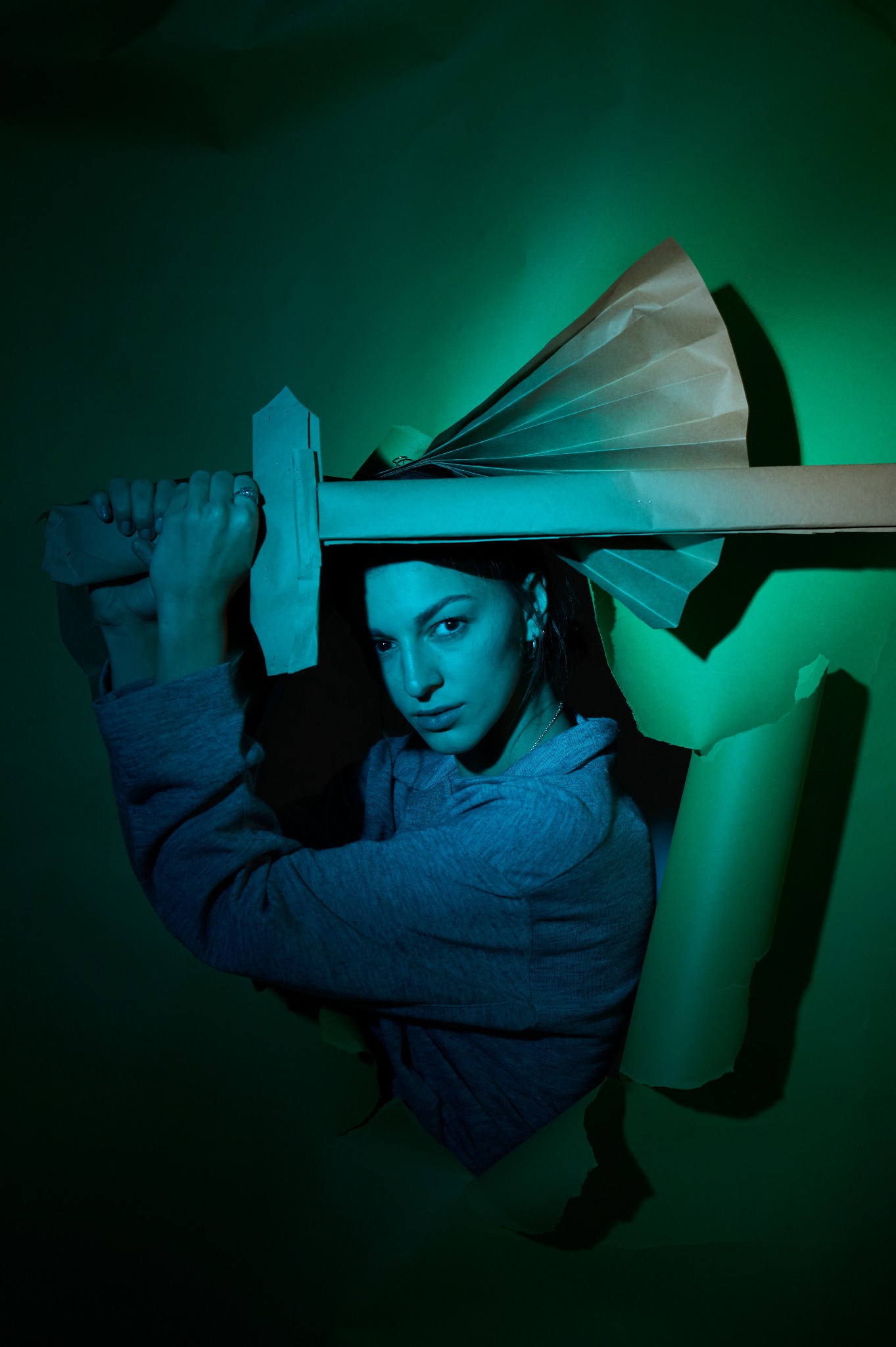 Młoda kobieta pozuje z tekturowym mieczem i wachlarzem, wystając z dziury w zielonym papierze, oświetlona niebiesko-zielonym światłem.