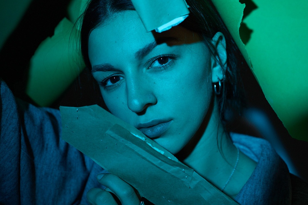 Portret kobiety o ciemnych włosach, w niebiesko-zielonym oświetleniu, z fragmentami podartego kartonu w tle i w dłoniach, patrzącej w obiektyw