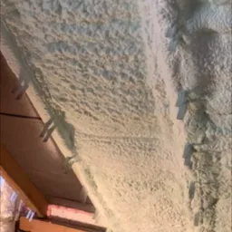 Poddasze po natrysku pianki poliuretanowej, widoczne krokwie i elementy mocujące, nierównomierna struktura piany.