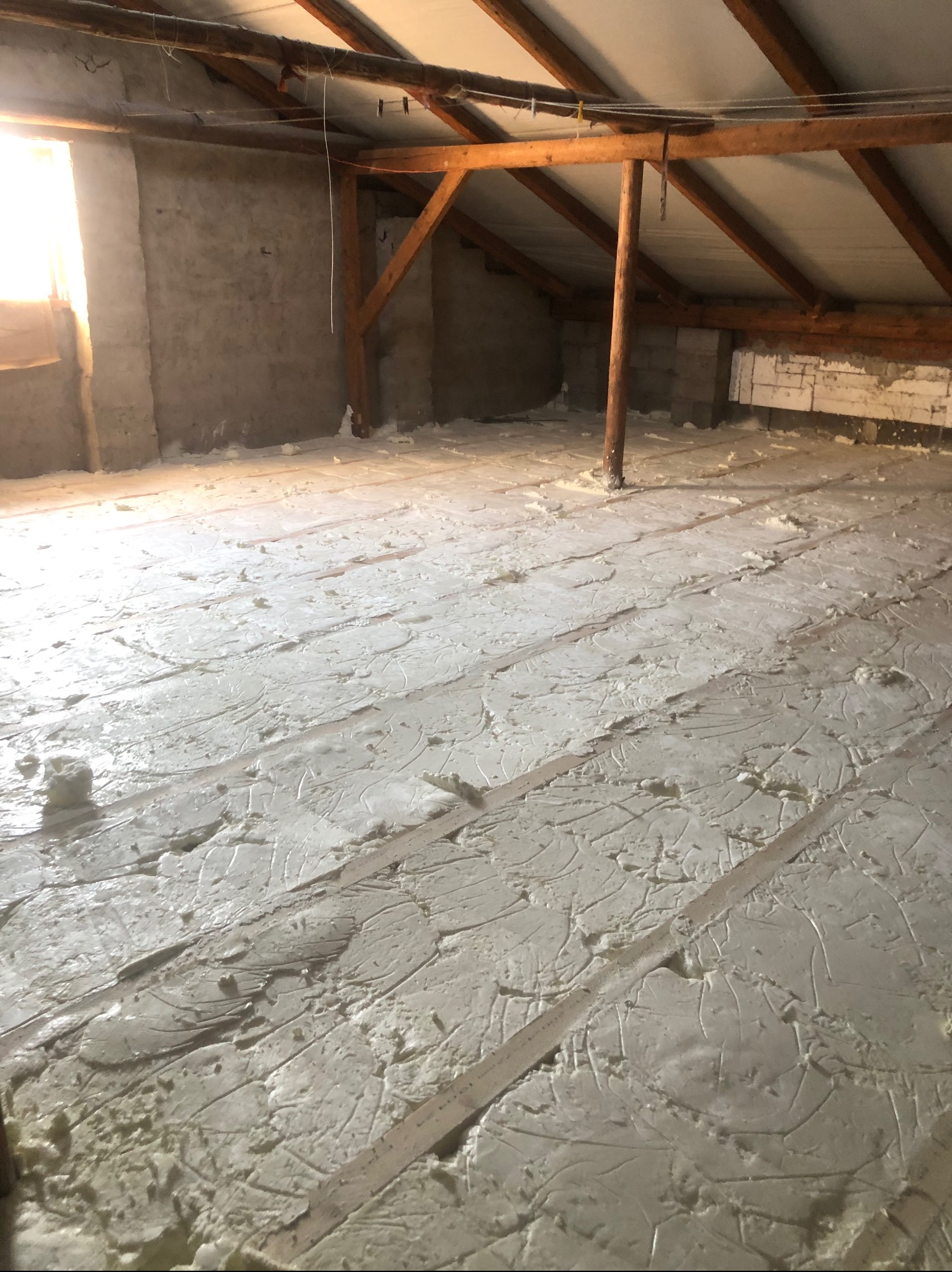 Poddasze z drewnianą konstrukcją dachu, po nałożeniu warstwy szarej piany izolacyjnej na podłogę, widoczne szczeliny między deskami.