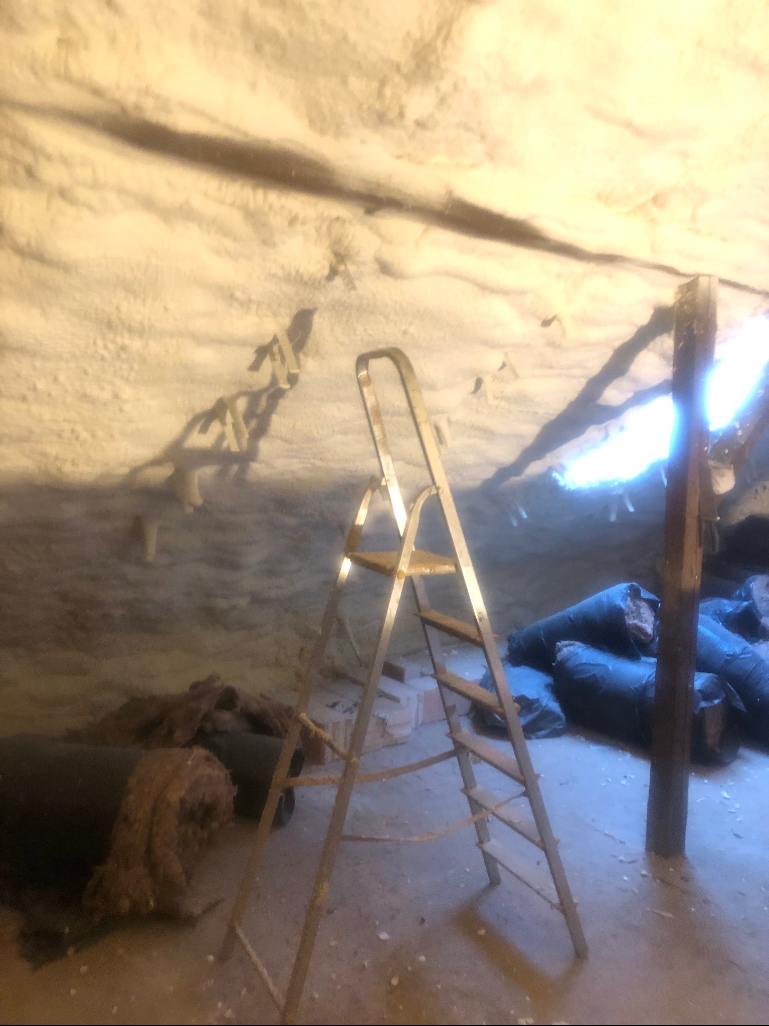 Poddasze w trakcie prac izolacyjnych, widoczna warstwa pianki na suficie, drabina aluminiowa, worki z materiałem izolacyjnym i fragment drewnianej konstrukcji dachu.