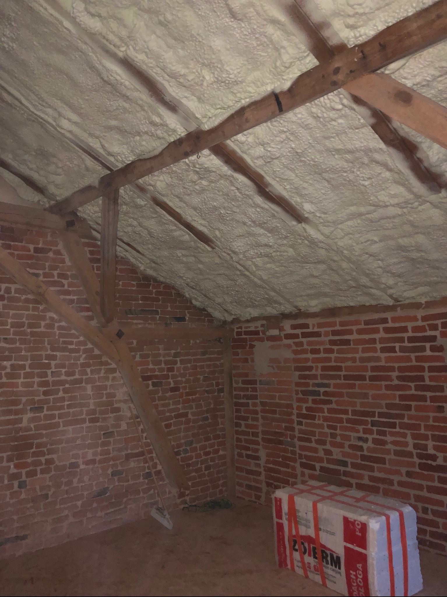 Poddasze z widoczną konstrukcją drewnianą i zaaplikowaną białą pianką izolacyjną, ceglana ściana, pakiet izolacji podłogowej w rogu pomieszczenia.