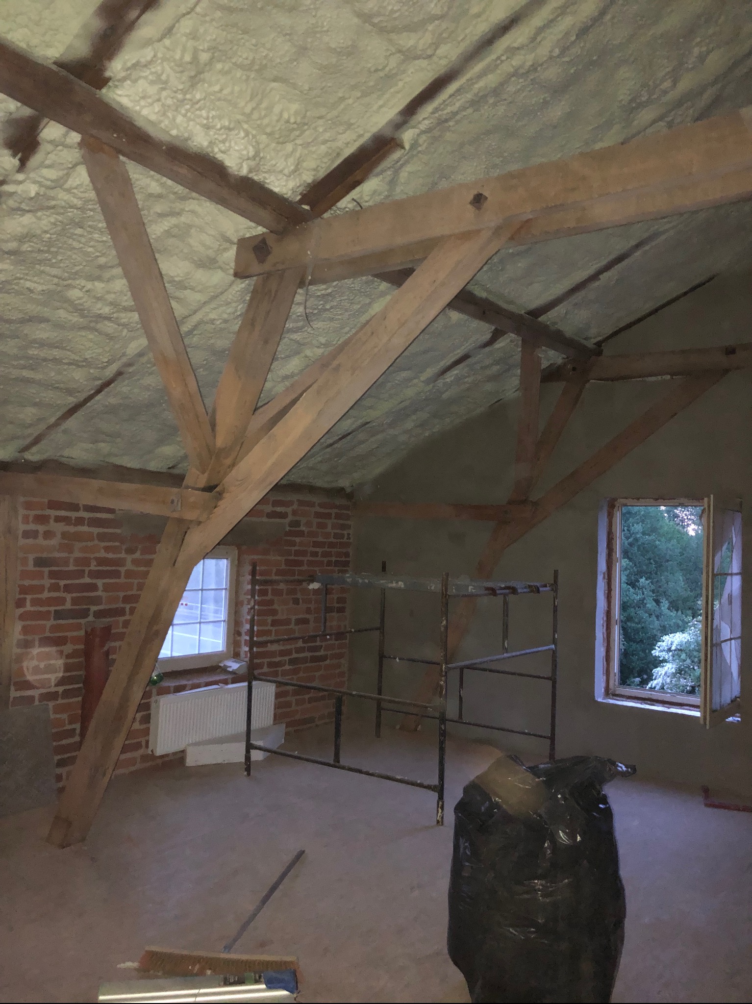 Poddasze z drewnianą konstrukcją dachu i zaizolowaną pianką sufitową, widoczna ściana z czerwonej cegły, okno z widokiem na zieleń, rusztowanie i worek na śmieci.