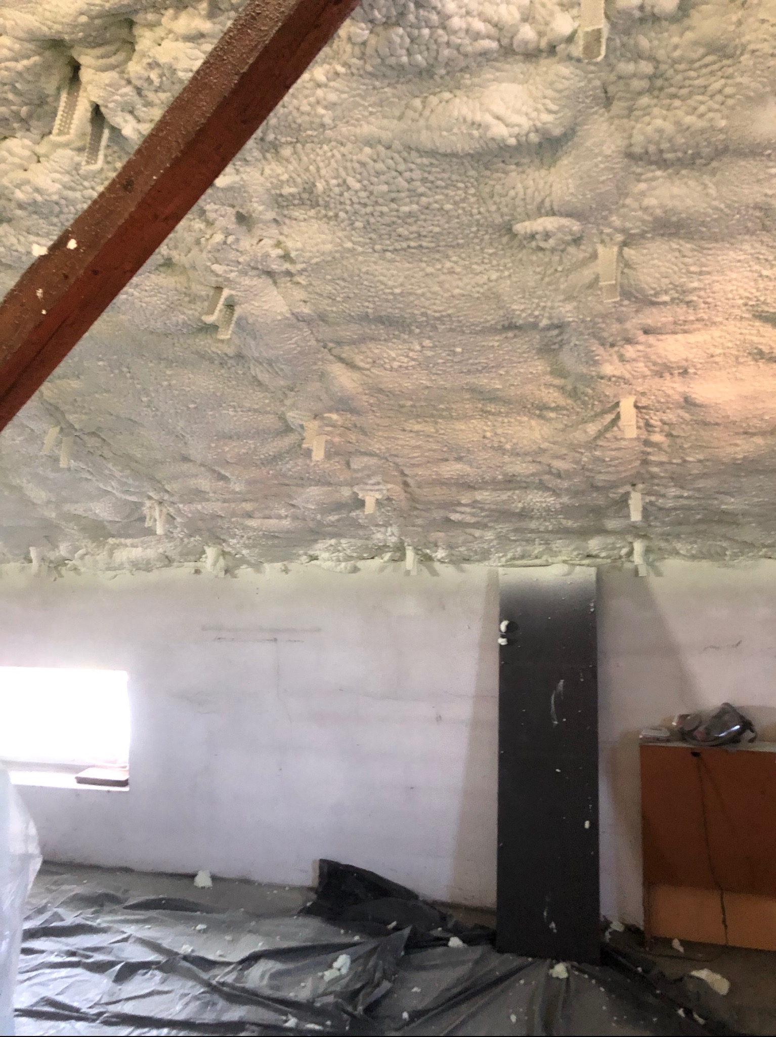 Poddasze z drewnianą belką i białą pianką izolacyjną, widoczne plastikowe uchwyty mocujące, podłoga zabezpieczona folią malarską.