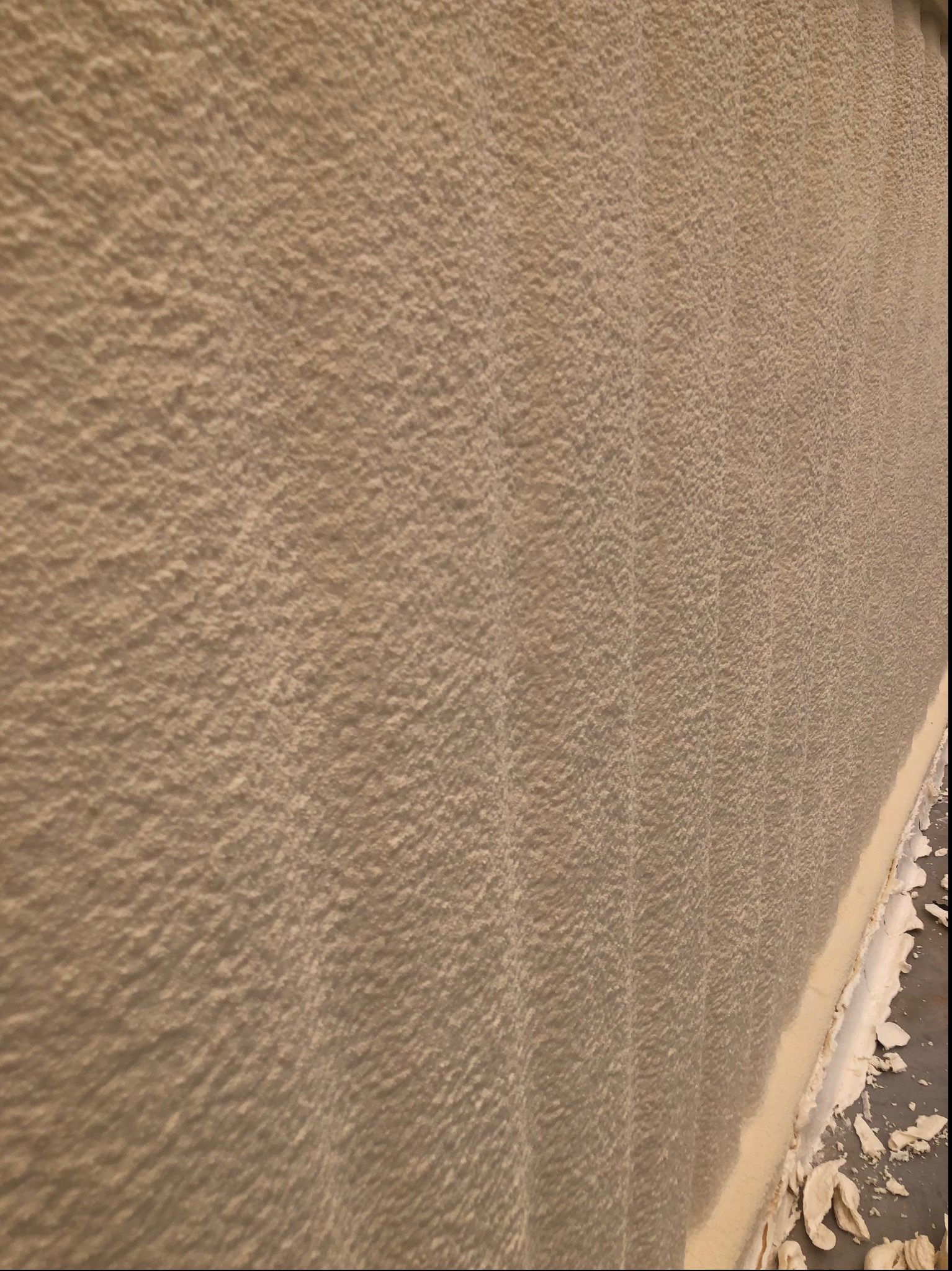 Ściana pokryta warstwą szarej pianki PUR o nieregularnej fakturze, z widocznymi fragmentami odpadającej pianki przy krawędzi podłogi. Ukazano detal wykończenia.