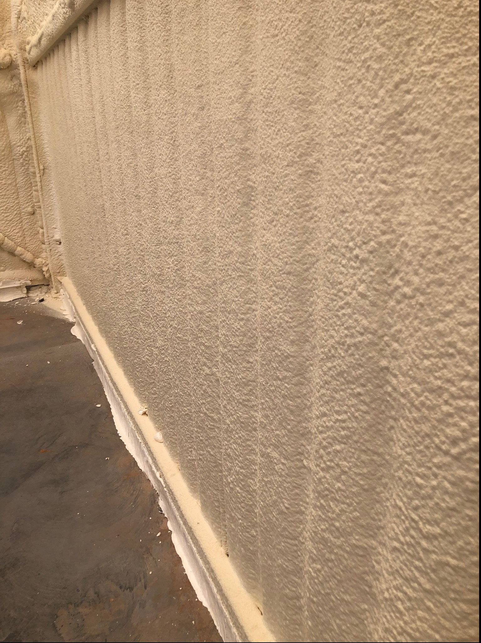 Pionowy kadr na ścianę pokrytą grubą warstwą pianki PUR, tworzącą nierówną, teksturowaną powierzchnię. Widoczne fragmenty instalacji i podłogi. Detal izolacji.