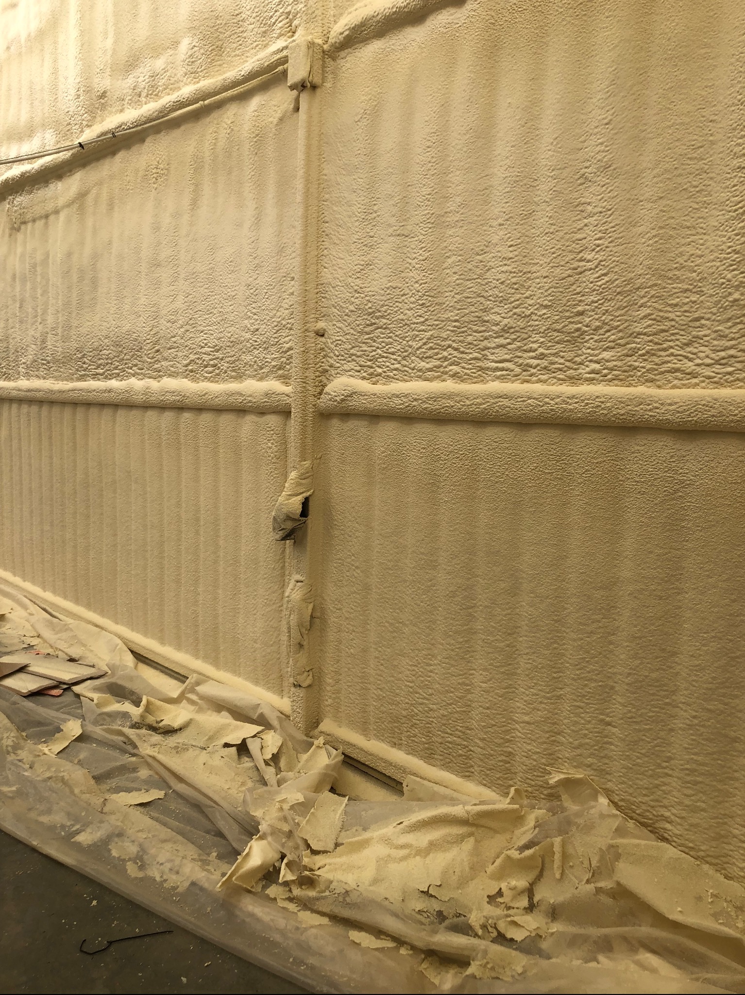 Pionowe ocieplenie ścian pianką PUR wewnątrz budynku, widoczne rury instalacyjne i zabezpieczenia podłogi folią. Struktura pianki gęsta, beżowa.