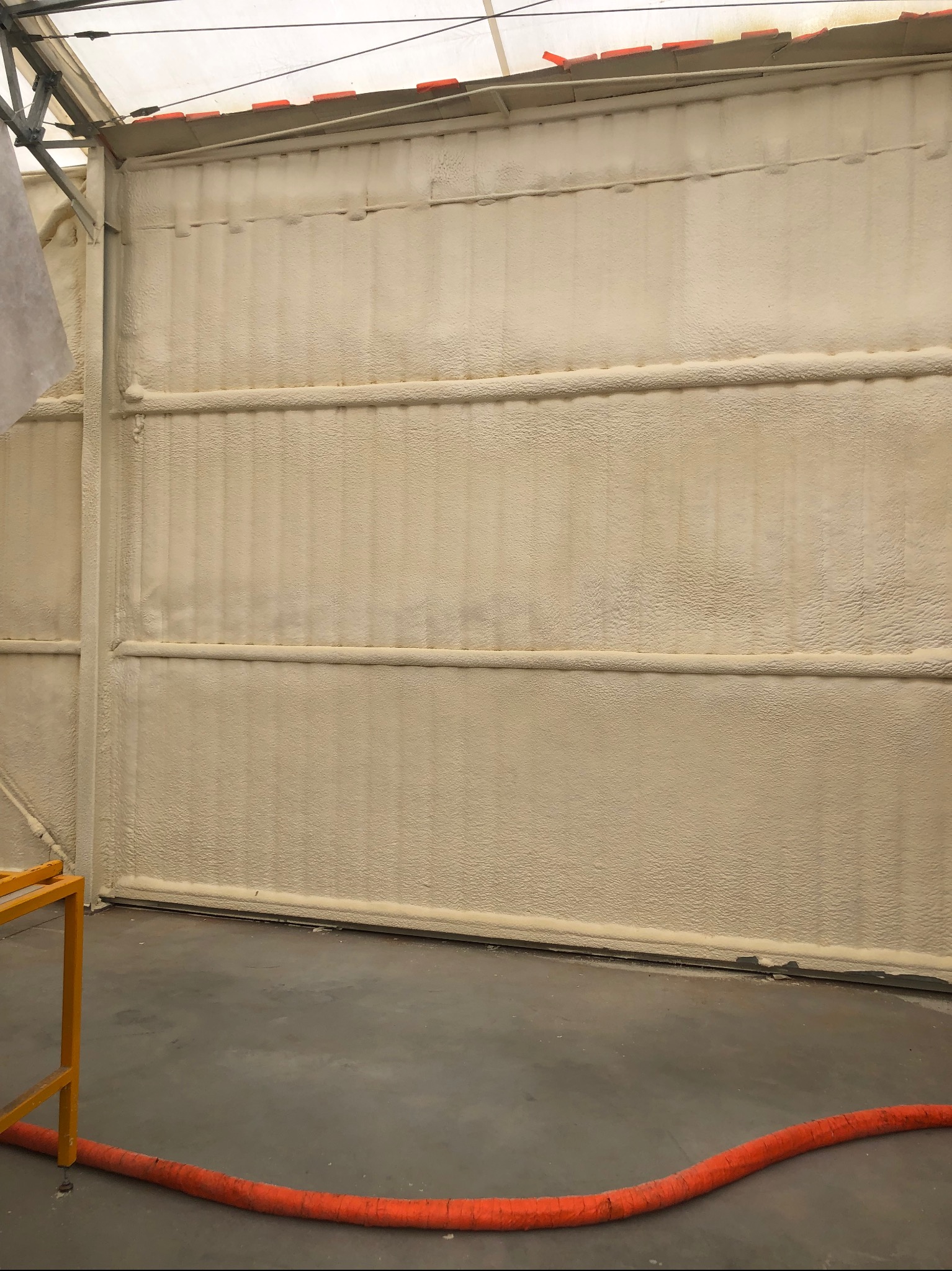 Ściana hali przemysłowej po ociepleniu pianką PUR, widoczne metalowe profile konstrukcyjne i pomarańczowy wąż do aplikacji pianki na posadzce.