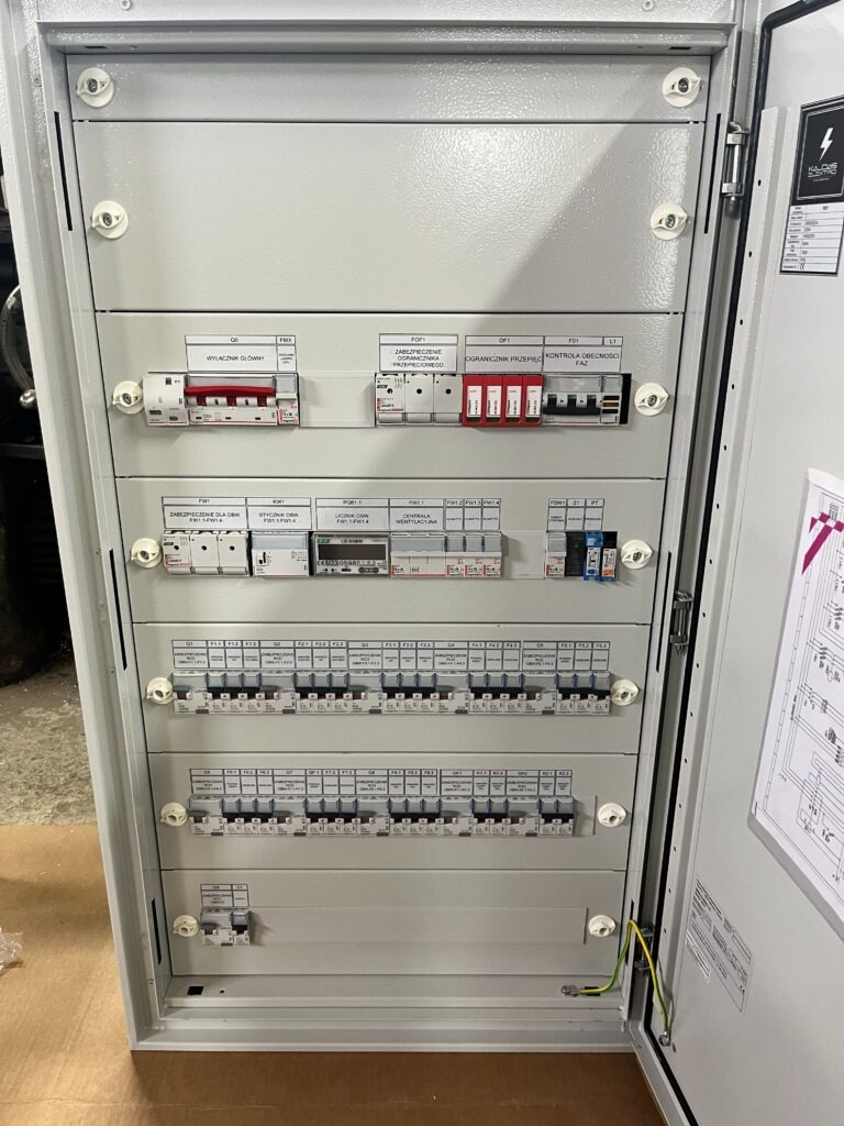 Otwarta, szara skrzynka elektryczna z bezpiecznikami, wyłącznikami i schematem na drzwiach. Widoczne przewody i elementy instalacji.