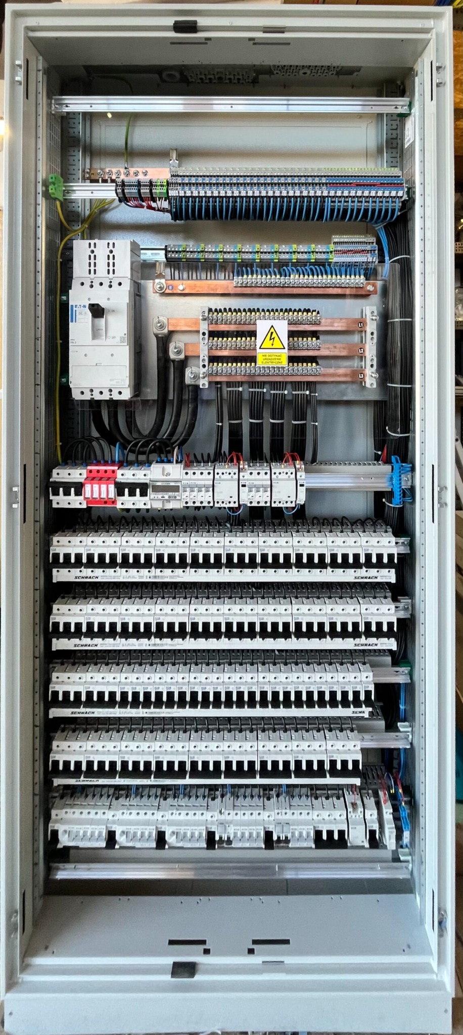 Rozdzielnica elektryczna z licznymi zabezpieczeniami, listwami i kablami. Widoczna tabliczka ostrzegawcza. Precyzyjny montaż w szarej obudowie.