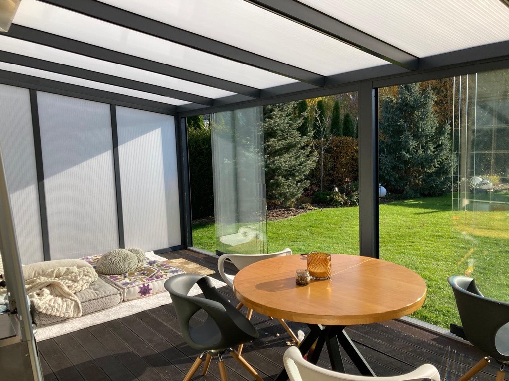 Nowoczesna konstrukcja z metalu i przezroczystych paneli tworząca zadaszone patio z widokiem na ogród. Wnętrze wyposażone w okrągły drewniany stół, krzesła i poduszki podłogowe.